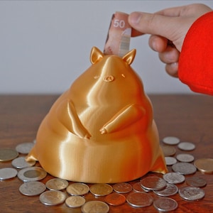 May include: A gold piggy bank shaped like a cartoon pig with a wide, round body and small legs. The piggy bank is sitting on a wooden table with coins scattered around it. A hand is putting a Canadian $50 bill into the piggy bank's slot.
