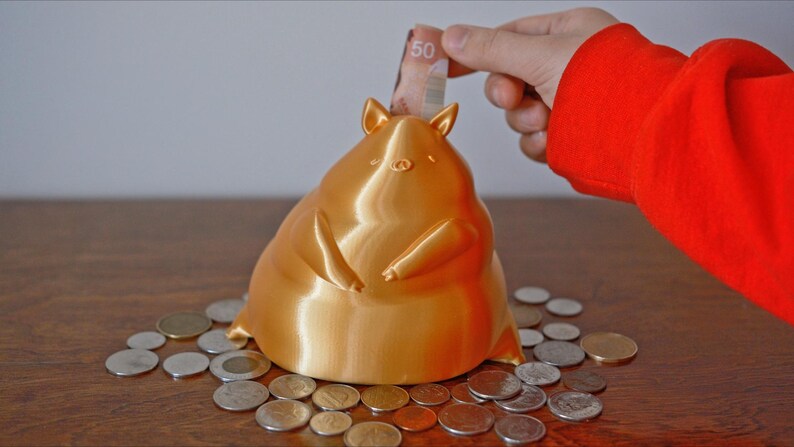 May include: A gold piggy bank shaped like a cartoon pig with a wide, round body and small legs. The piggy bank is sitting on a wooden table with coins scattered around it. A hand is putting a Canadian $50 bill into the piggy bank's slot.
