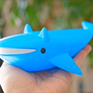 May include: A bright blue whale figurine with a white mouth and black eye is held in a hand. The whale has fins and a tail. The figurine is a toy or decorative item.