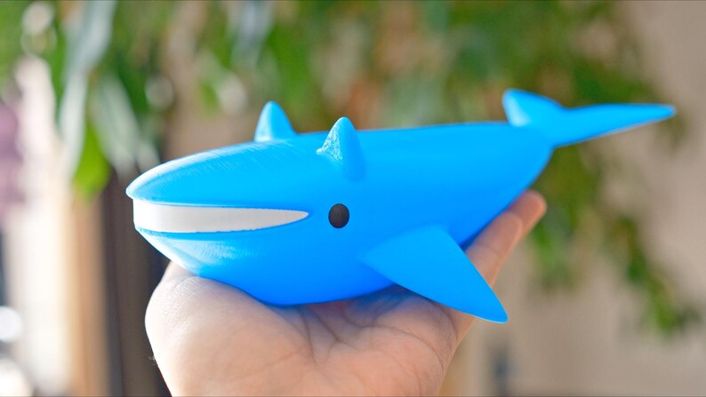 May include: A bright blue whale figurine with a white mouth and black eye is held in a hand. The whale has fins and a tail. The figurine is a toy or decorative item.