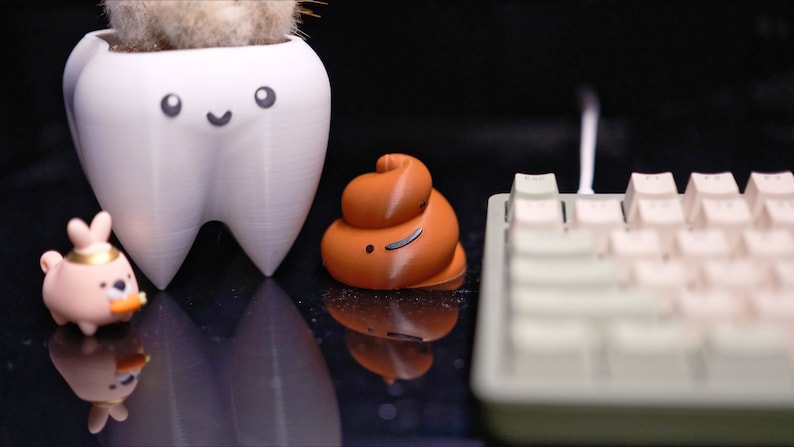 May include: A white tooth-shaped planter with a smiling face, a brown poop-shaped figurine with a smiley face, a pink bunny figurine holding a carrot, and a white keyboard with beige keys.