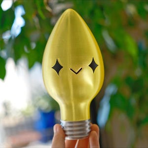 May include: A gold-coloured lightbulb-shaped object with a smiling face cut out of the front. The face has two diamond-shaped eyes and a small, downward-pointing mouth. The base of the bulb is silver. The background is blurred green foliage.