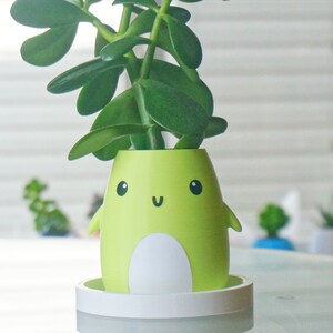 Puede incluir: Una maceta de cer&aacute;mica verde y blanca con forma de personaje de dibujos animados con una cara sonriente. La maceta tiene un platillo blanco y est&aacute; llena de una planta verde.