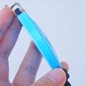 Puede incluir: Un llavero azul claro con un anillo de metal plateado y un cord&oacute;n negro.