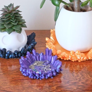May include: Three decorative crystal-shaped plant saucers in blue, purple, and orange. The saucers are made of a 3D printed material and have a unique, geometric design. The purple saucer is filled with jewellery.