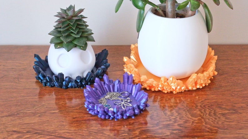 May include: Three decorative crystal-shaped plant saucers in blue, purple, and orange. The saucers are made of a 3D printed material and have a unique, geometric design. The purple saucer is filled with jewellery.