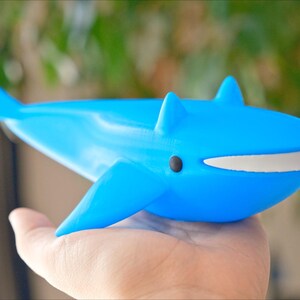 May include: A bright blue whale figurine with a white mouth and black eye. The toy whale has fins and a tail, and is held in a hand. The background is blurred with green foliage.