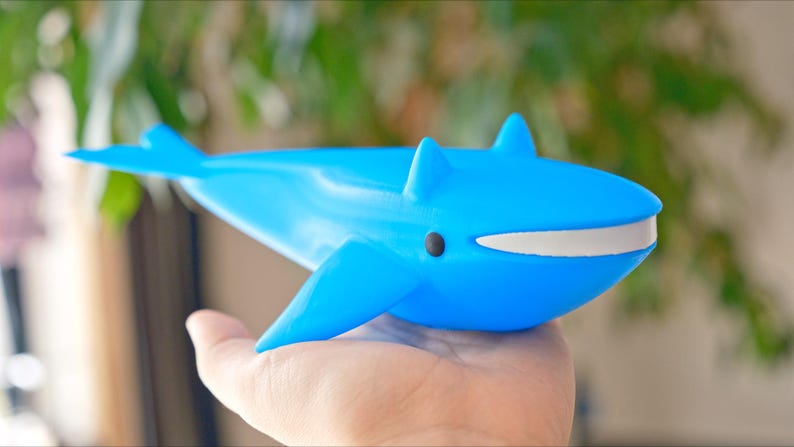 May include: A bright blue whale figurine with a white mouth and black eye. The toy whale has fins and a tail, and is held in a hand. The background is blurred with green foliage.