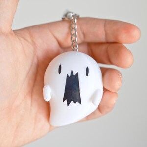 May include: A white ghost-shaped keychain with a black mouth and small black eyes. The keychain is attached to a silver chain. The ghost has small arms and a rounded body. The background is a plain, light color.