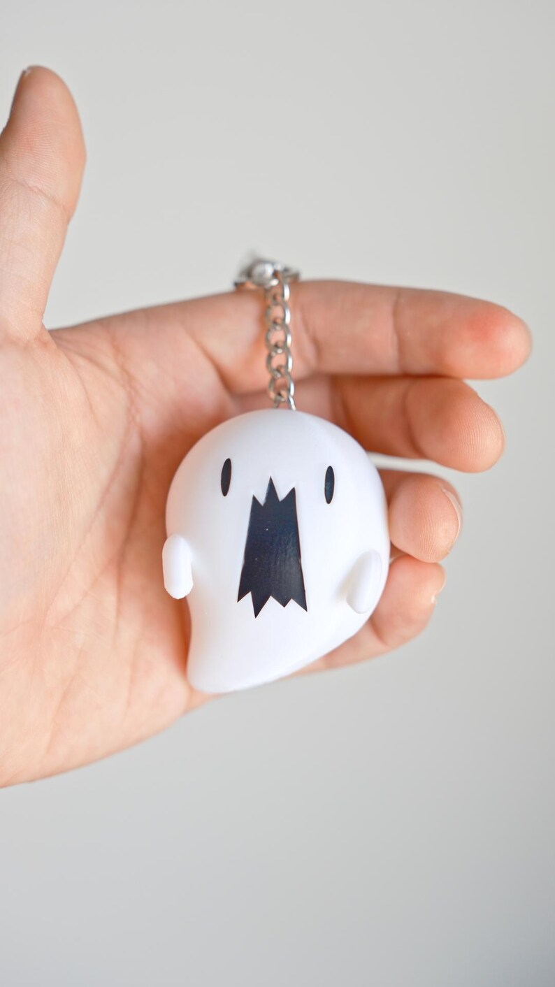 May include: A white ghost-shaped keychain with a black mouth and small black eyes. The keychain is attached to a silver chain. The ghost has small arms and a rounded body. The background is a plain, light color.