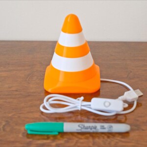 May include: An orange and white traffic cone-shaped lamp with a USB cable and on/off switch. A green-capped Sharpie marker is below the lamp. The lamp is on a wooden surface.