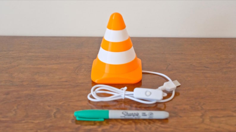 May include: An orange and white traffic cone-shaped lamp with a USB cable and on/off switch. A green-capped Sharpie marker is below the lamp. The lamp is on a wooden surface.