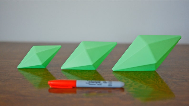 May include: Three green diamond-shaped objects of varying sizes, arranged in a row from smallest to largest on a wooden surface.