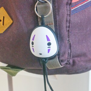 May include: A backpack with a No-Face keychain and a Totoro pin. The No-Face keychain is white with purple markings and black details. The Totoro pin is round with a blue background and a smiling Totoro character. The backpack is dark purple.
