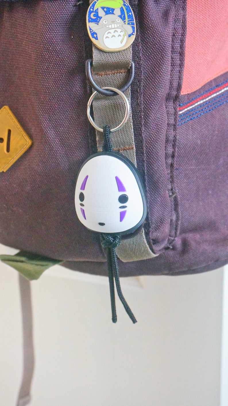 May include: A backpack with a No-Face keychain and a Totoro pin. The No-Face keychain is white with purple markings and black details. The Totoro pin is round with a blue background and a smiling Totoro character. The backpack is dark purple.