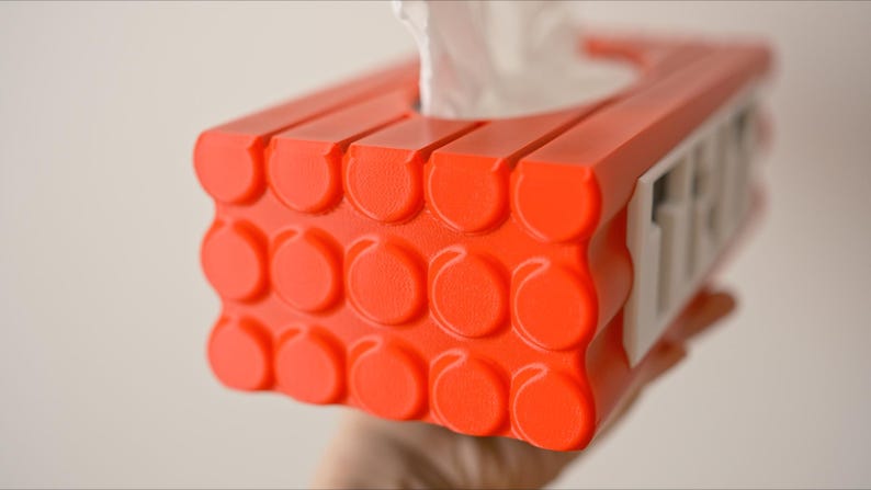 May include: Orange tissue box holder with a white label that says "FREE". The holder is designed to look like a stack of building blocks.