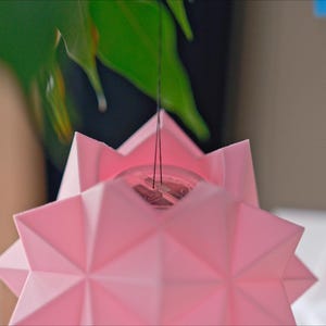 Pode incluir: Um abajur de origami em forma de estrela rosa pendurado em um cord&atilde;o. O abajur &eacute; feito de papel e tem um design geom&eacute;trico.