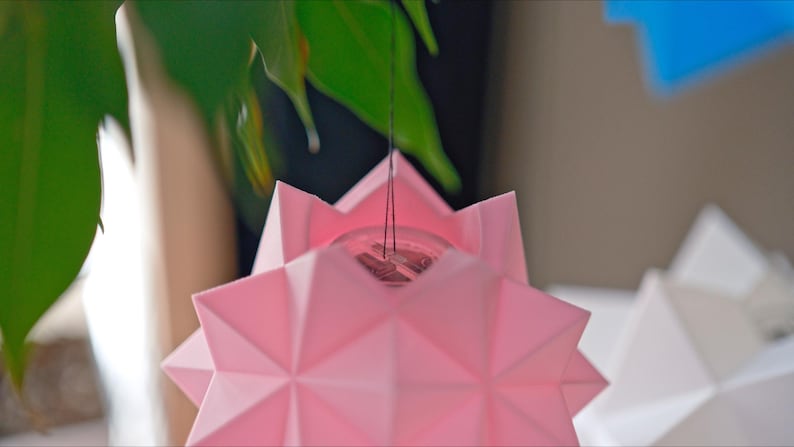 Pode incluir: Um abajur de origami em forma de estrela rosa pendurado em um cord&atilde;o. O abajur &eacute; feito de papel e tem um design geom&eacute;trico.