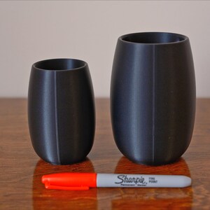 May include: Two black cylindrical cups, one taller than the other, on a wooden surface. A red and white marker is placed between the cups.