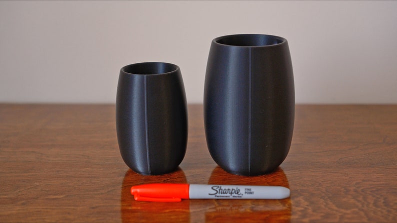 May include: Two black cylindrical cups, one taller than the other, on a wooden surface. A red and white marker is placed between the cups.