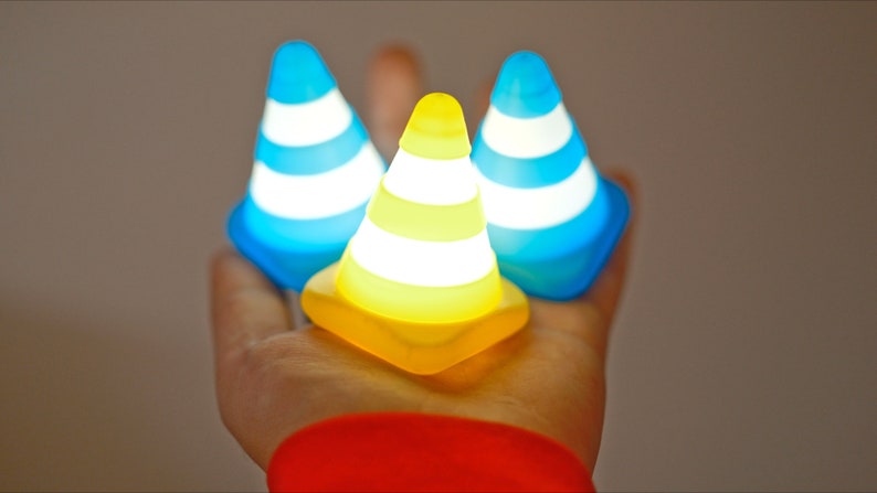 Op de afbeelding: Drie kleine, kegelvormige, lichtgevende speelgoedjes. Twee zijn blauw met witte strepen en &eacute;&eacute;n is geel met witte strepen.