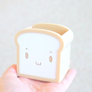May include: A white and beige bread-shaped pencil holder with a cute cartoon face.