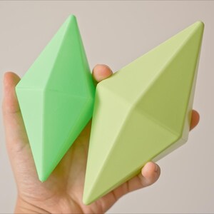 May include: Two green diamond-shaped toys made of a soft, pliable material. The toys are different shades of green and are held in a person's hand.