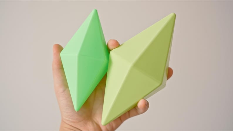 May include: Two green diamond-shaped toys made of a soft, pliable material. The toys are different shades of green and are held in a person's hand.