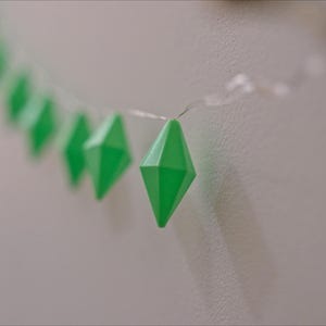 May include: A string of bright green, diamond-shaped decorative lights. The lights are strung on a thin, clear wire and are set against a neutral-colored wall. The geometric shapes are evenly spaced.