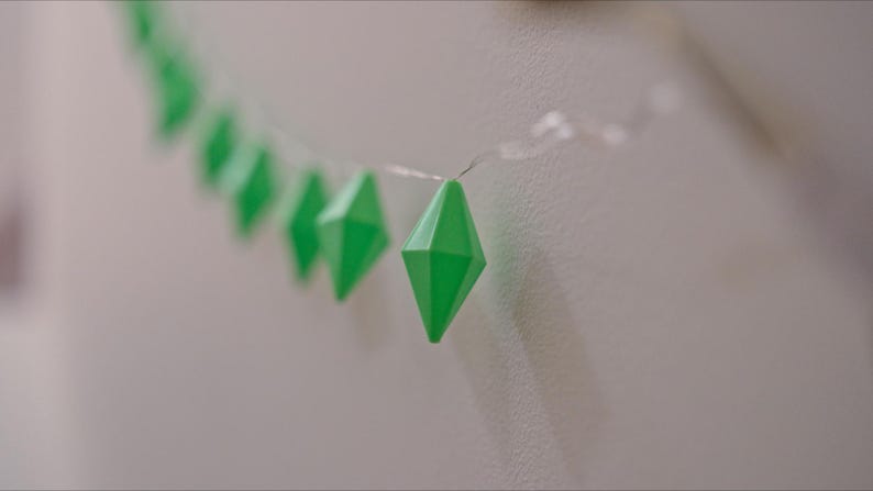 May include: A string of bright green, diamond-shaped decorative lights. The lights are strung on a thin, clear wire and are set against a neutral-colored wall. The geometric shapes are evenly spaced.