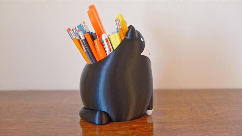 May include: A black cat-shaped desk organizer filled with pencils, pens, and markers. The cat has white accents on its face and paws. The organizer is sitting on a wooden surface.