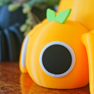 May include: Close-up of two whimsical, pumpkin-shaped objects. One is black, the other is bright orange, both with large, round, black eyes outlined in white. Each has a green leaf on top and curved, yellow appendages. The objects are on a wooden surface.