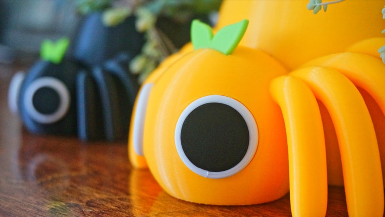 May include: Close-up of two whimsical, pumpkin-shaped objects. One is black, the other is bright orange, both with large, round, black eyes outlined in white. Each has a green leaf on top and curved, yellow appendages. The objects are on a wooden surface.