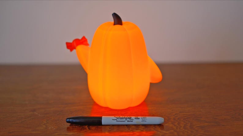 May include: A bright orange pumpkin-shaped night light with a red bow on the side. The light is on and glowing.