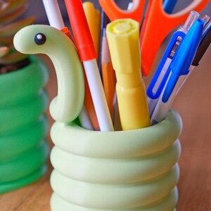 May include: A light green, snake-shaped desk organizer filled with pens, pencils, and scissors. The organizer is made of a smooth, coiled material. A second, similar organizer is visible in the background. The desk organizer is a fun and functional item for any desk.