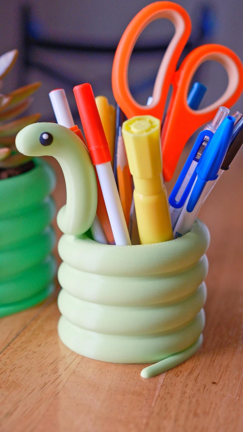 May include: A light green, snake-shaped desk organizer filled with pens, pencils, and scissors. The organizer is made of a smooth, coiled material. A second, similar organizer is visible in the background. The desk organizer is a fun and functional item for any desk.