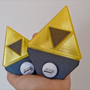 May include: Two gold-colored Triforce-shaped USB charging adapters with black bases. The Triforce symbol is a popular icon from the Legend of Zelda video game series.