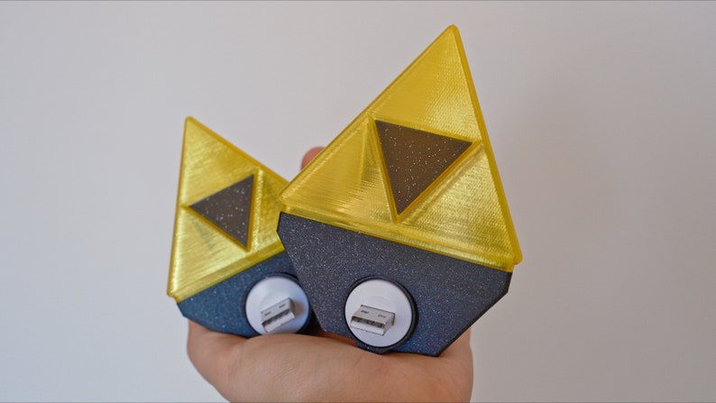 May include: Two gold-colored Triforce-shaped USB charging adapters with black bases. The Triforce symbol is a popular icon from the Legend of Zelda video game series.