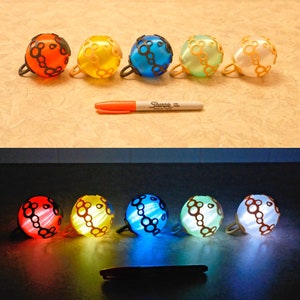 May include: A collection of five colorful, spherical bells with black handles and decorative patterns. The bells are in shades of orange, yellow, blue, green, and white. A Sharpie marker is placed next to the bells.