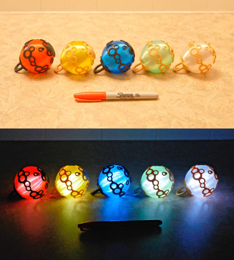 May include: A collection of five colorful, spherical bells with black handles and decorative patterns. The bells are in shades of orange, yellow, blue, green, and white. A Sharpie marker is placed next to the bells.