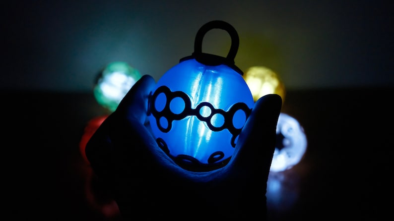 May include: A glowing blue ornament with a black decorative design is held in a hand. Other colorful, glowing ornaments are visible in the background. The ornament has a loop at the top for hanging. The overall theme is festive and decorative.
