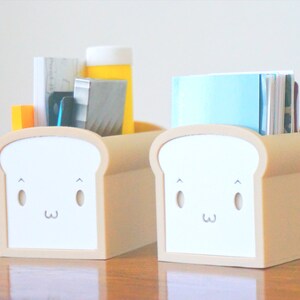 May include: Two beige bread-shaped desk organizers with cute cartoon faces. One organizer holds a stack of papers, a ruler, a pen, and a yellow bottle. The other organizer holds a stack of blue and white papers.