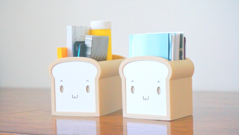 May include: Two beige bread-shaped desk organizers with cute cartoon faces. One organizer holds a stack of papers, a ruler, a pen, and a yellow bottle. The other organizer holds a stack of blue and white papers.