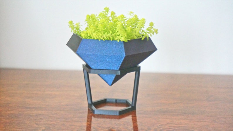 May include: A blue diamond-shaped planter with a green succulent plant inside, sitting on a black metal stand. The planter has a sparkly finish.