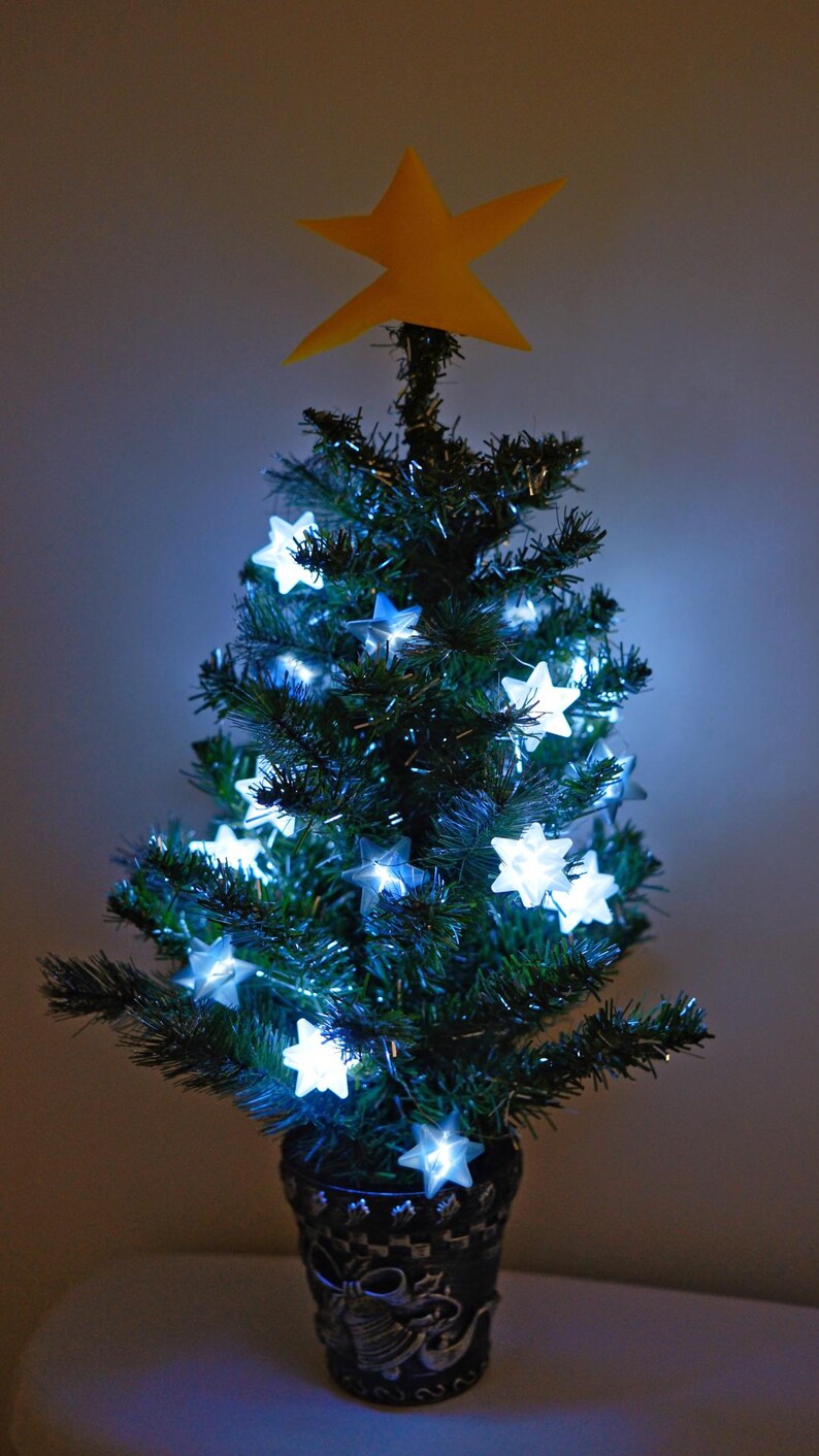 May include: A small artificial Christmas tree decorated with white star-shaped lights and a yellow paper star on top. The tree is in a black and silver planter.