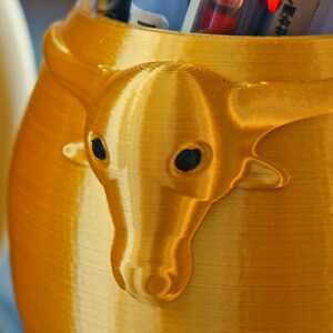 Puede incluir: Un contenedor de color dorado, impreso en 3D, con un dise&ntilde;o de cabeza de toro de color dorado, impreso en 3D. El contenedor est&aacute; dise&ntilde;ado para guardar bol&iacute;grafos o l&aacute;pices.