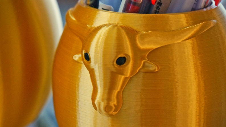 Puede incluir: Un contenedor de color dorado, impreso en 3D, con un dise&ntilde;o de cabeza de toro de color dorado, impreso en 3D. El contenedor est&aacute; dise&ntilde;ado para guardar bol&iacute;grafos o l&aacute;pices.