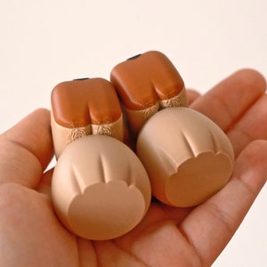 May include: Two small, brown and beige ceramic figurines with rounded shapes. The figurines are shaped like the bottom half of a person, with brown legs and beige rounded bottoms.
