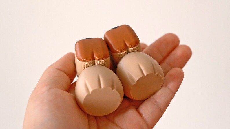 May include: Two small, brown and beige ceramic figurines with rounded shapes. The figurines are shaped like the bottom half of a person, with brown legs and beige rounded bottoms.