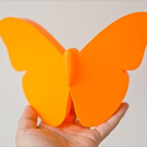 May include: A bright orange butterfly decoration is held in a hand. The butterfly has large, symmetrical wings and a simple, modern design. The vibrant color makes it a striking decorative piece.